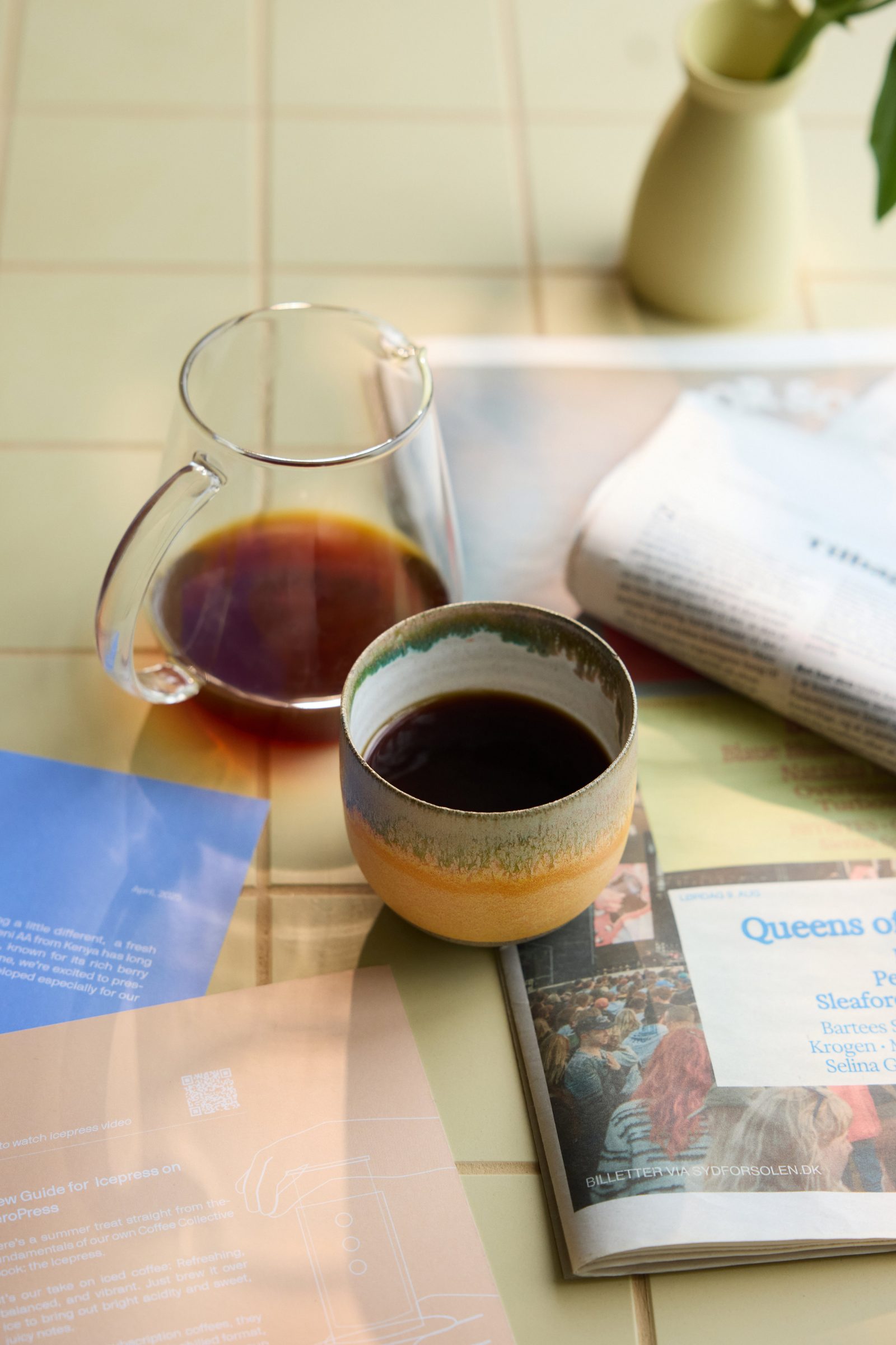 Coffee cup and glass carafe on a tiled counter, with a magazine underneath